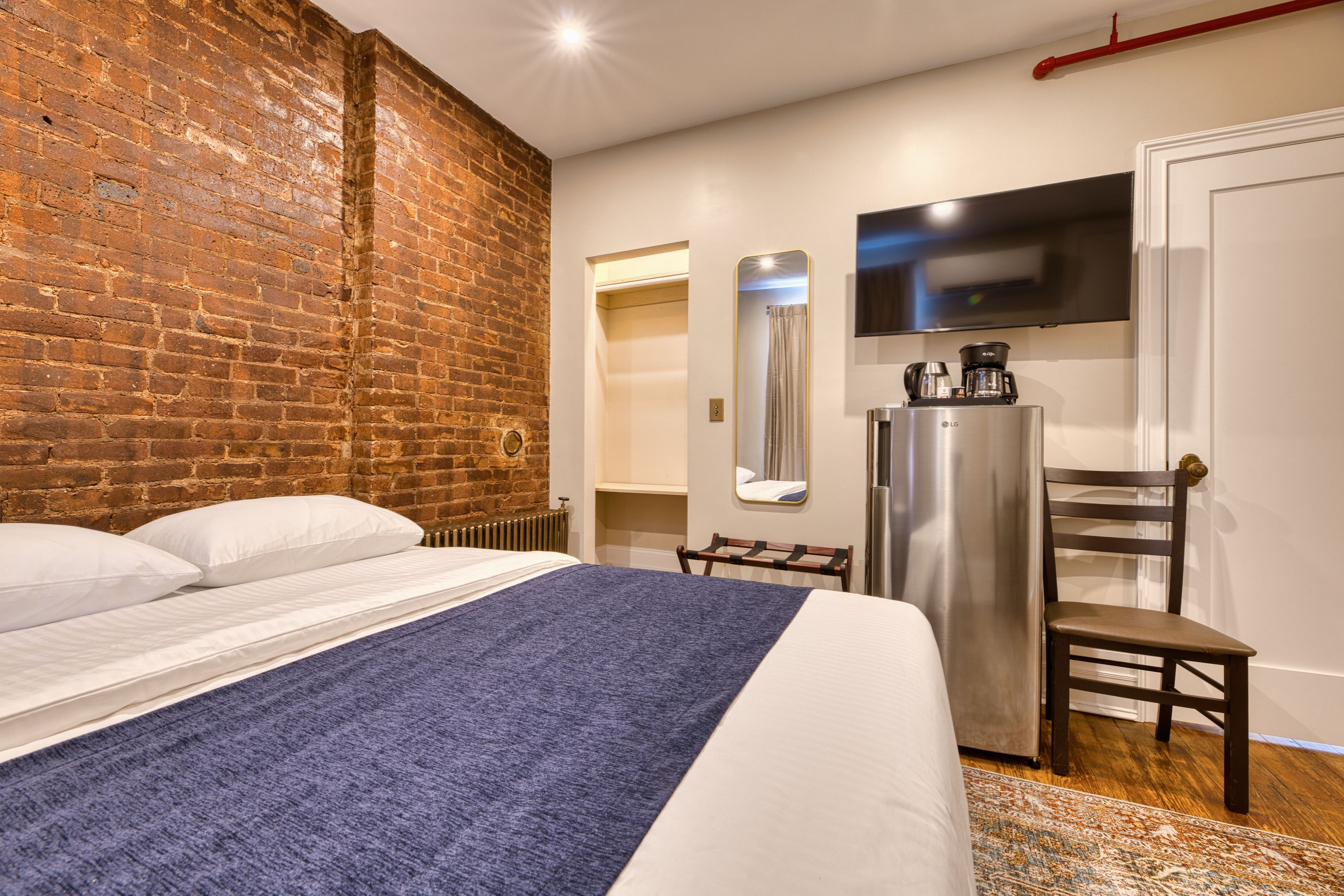 Habitación de hotel con pared de ladrillo, cama con ropa blanca, manta azul, televisor de pantalla plana, cafetera y una pequeña silla de madera.