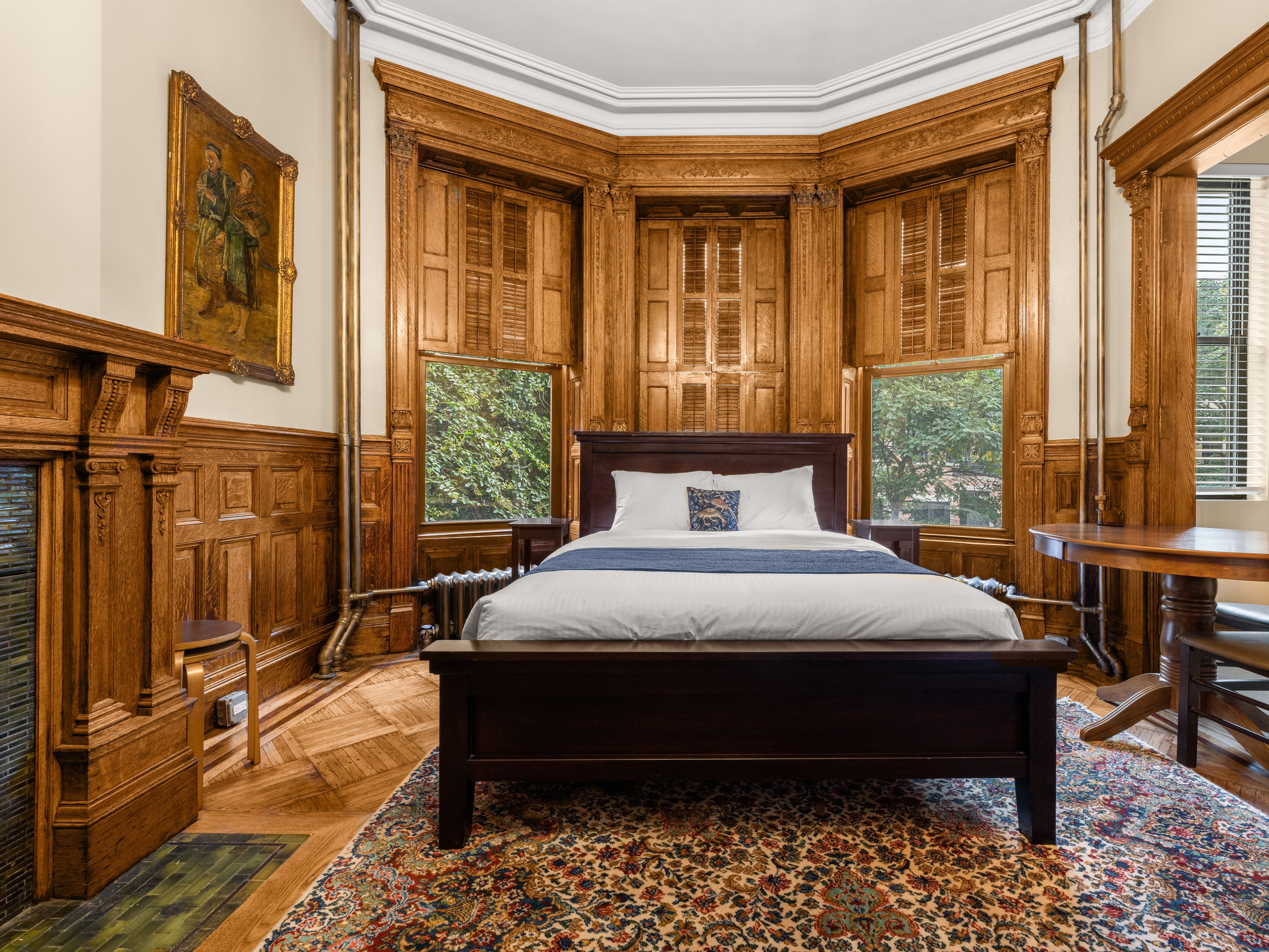 A traditional bedroom with a large bed, wooden paneling, and a view of greenery through the windows.