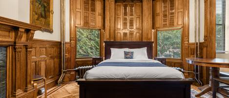 A traditional bedroom with a large bed, wooden paneling, and a view of greenery through the windows.