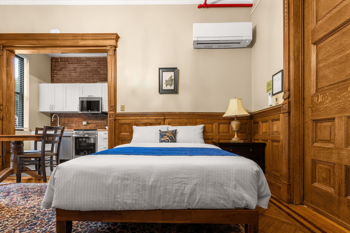A hotel room with a wooden bed, a kitchen area with white cabinets, and a brick wall.