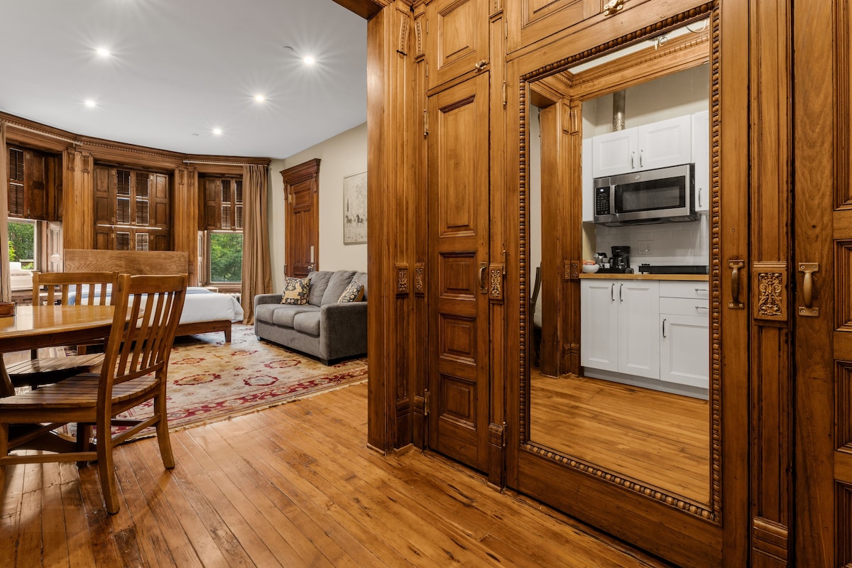 A spacious room with wooden flooring, a dining table with chairs, a sofa, and a kitchen area with white cabinets and a microwave.