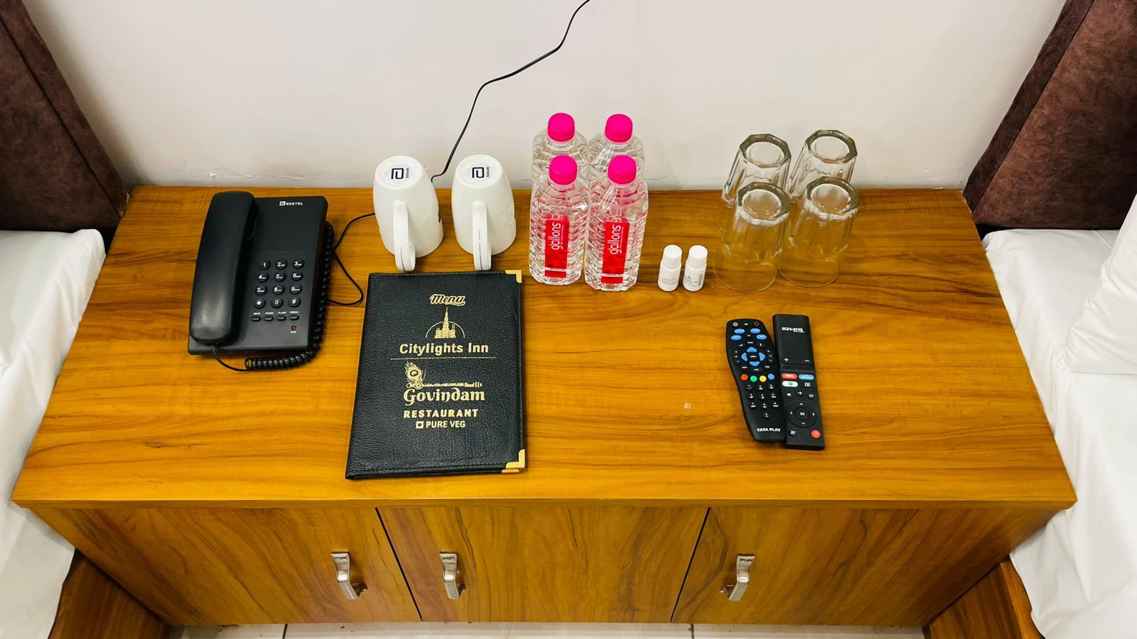 A wooden table with a black telephone, two white water bottles, a black menu book, a pair of glasses, and a remote control.