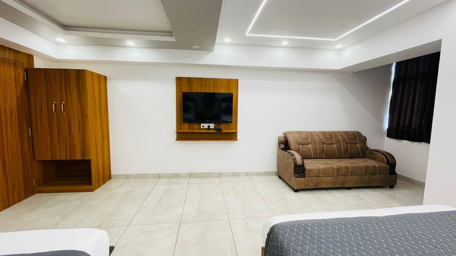 A modern bedroom with a bed, a wooden wardrobe, a sofa, and a TV mounted on the wall.