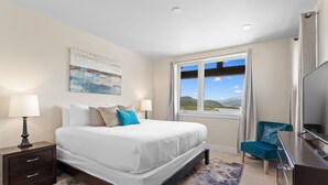 A bedroom with a large bed, bedside lamps, a desk, a chair, a TV, a window with a view, and a painting on the wall. - New 3BR Condo Within Walking Distance to Marina and Mins From Ski Resorts! (Dillon)