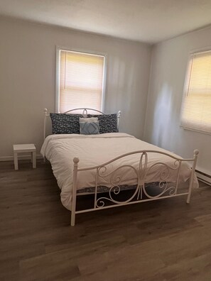 Une chambre avec un lit simple, une petite table de chevet et deux fenêtres équipées de stores.