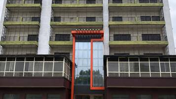 A multi-story building with a central glass entrance, balconies on each floor, and a brick-paved ground.