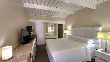 Una habitación de hotel con cama, lámparas de noche, escritorio con lámpara, televisor y espejo.