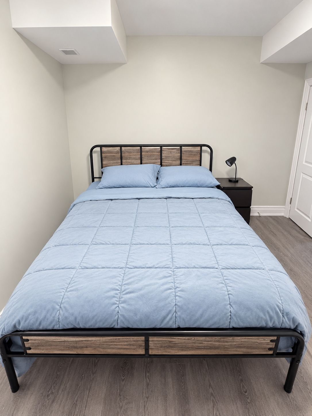 A bed with a wooden frame and a blue quilt, a bedside table with a lamp, and a door in the background.