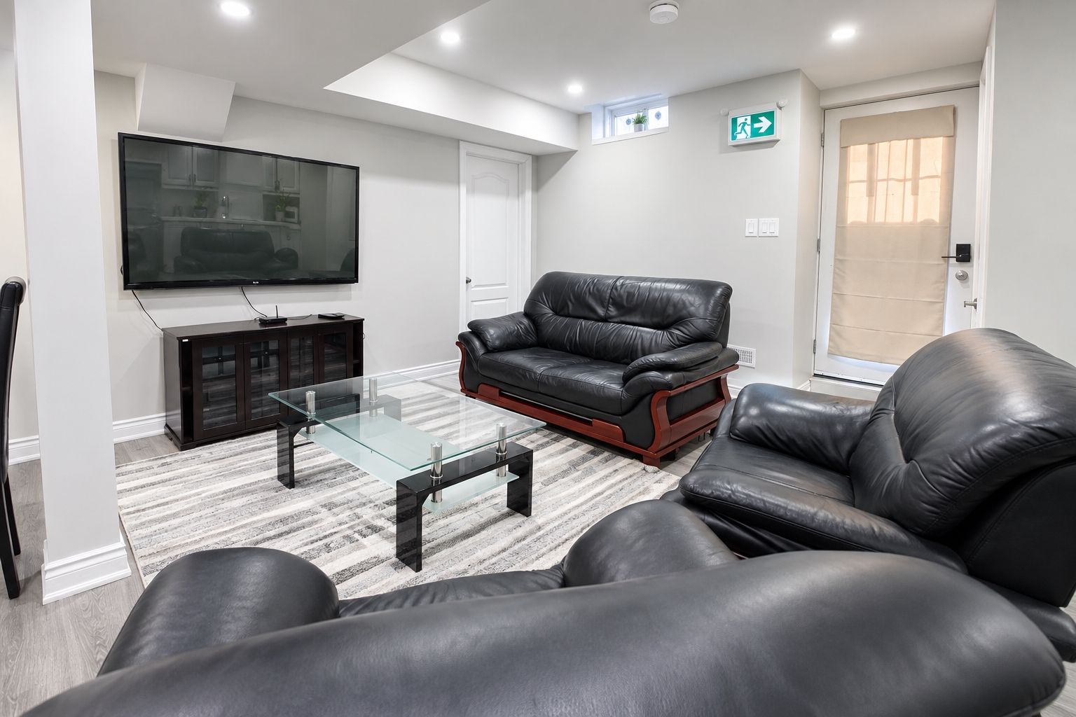 A modern living room with a glass coffee table, leather sofas, a flat-screen TV, and a door with an emergency exit sign.