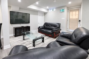 A modern living room with a glass coffee table, leather sofas, a flat-screen TV, and a door with an emergency exit sign.