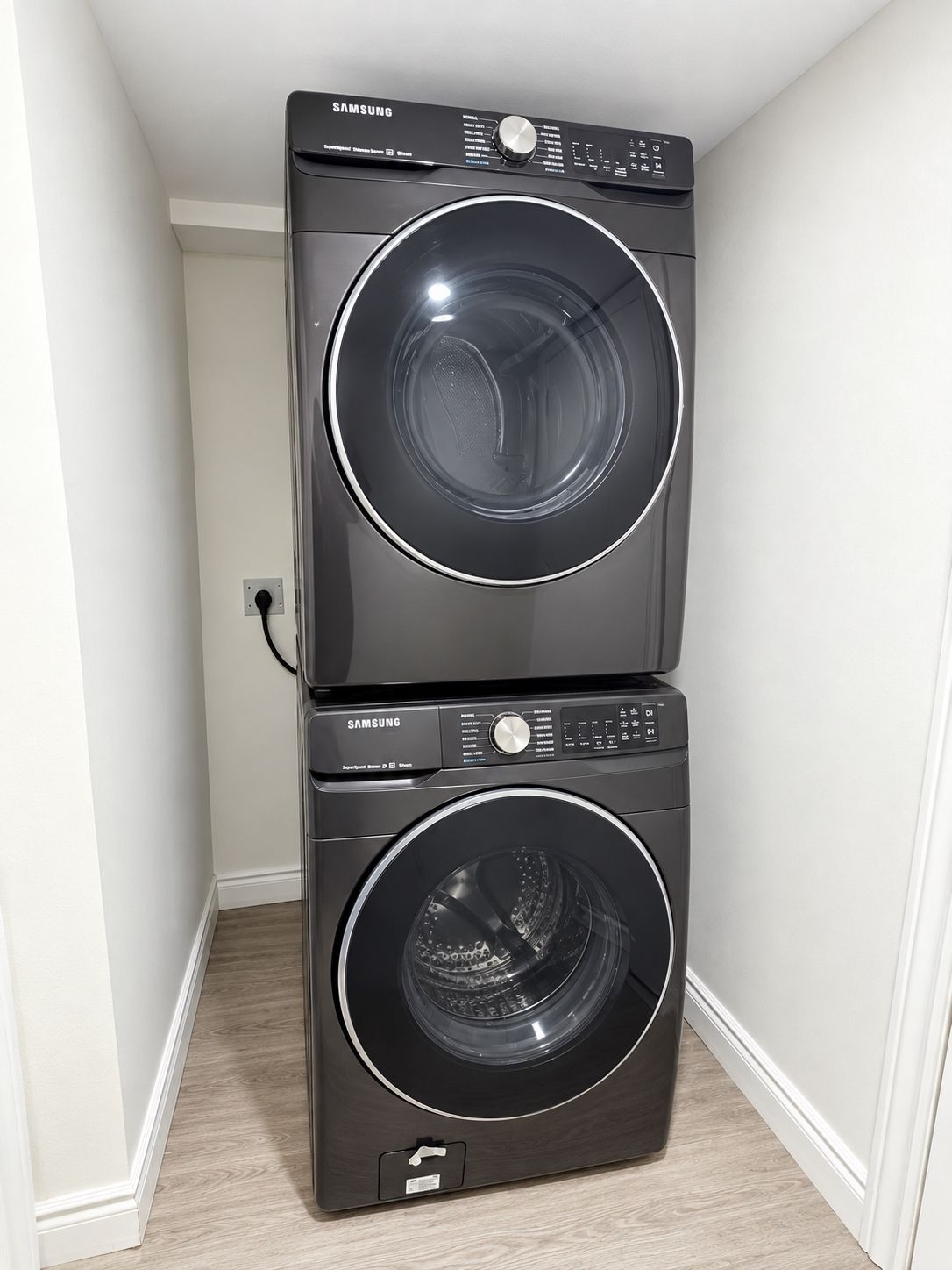 A Samsung stacked washer and dryer unit in a corner of a room with white walls and wooden flooring.