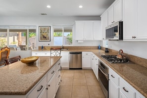 A modern kitchen with white cabinets, granite countertops, stainless steel appliances, and a large window providing natural light. - Saltwater pool + outdoor movies (Indio)