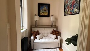A narrow hallway with a white sofa, two lamps, a radiator, a patterned rug, and a painting on the wall.