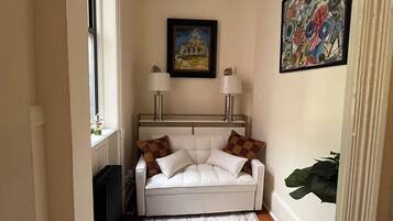 A narrow hallway with a white sofa, two lamps, a radiator, a patterned rug, and a painting on the wall.