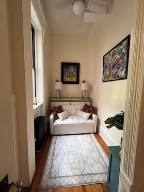 A narrow hallway with a white sofa, two lamps, a radiator, a patterned rug, and a painting on the wall.