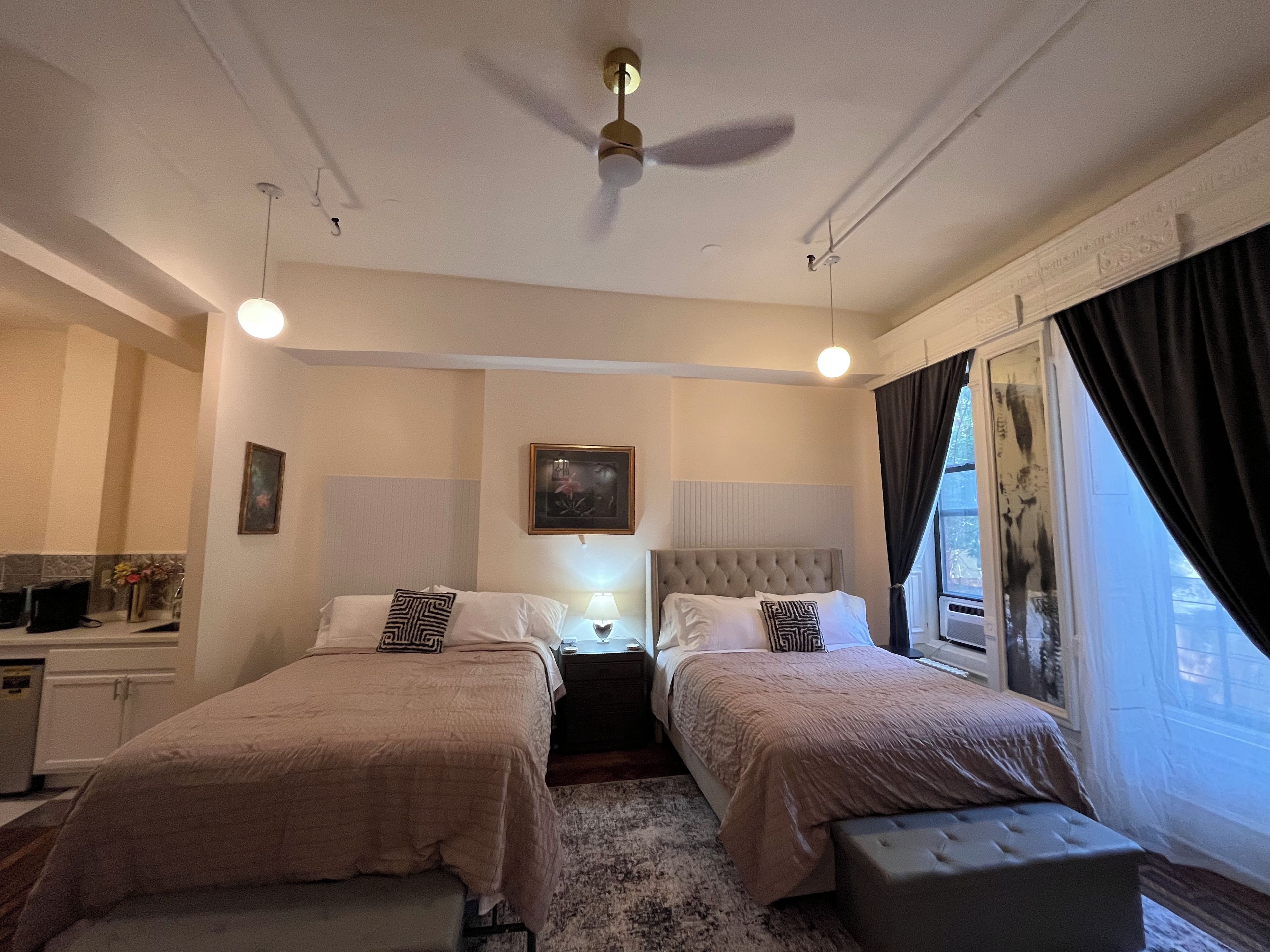 A hotel room with two beds, a ceiling fan, a bedside table with a lamp, a window with curtains, and a painting on the wall.
