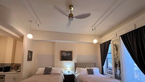 Habitación de hotel con dos camas, un ventilador de techo, una mesita de noche con lámpara, una ventana con cortinas y un cuadro en la pared.