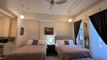 A hotel room with two beds, a ceiling fan, a bedside table with a lamp, a window with curtains, and a painting on the wall.