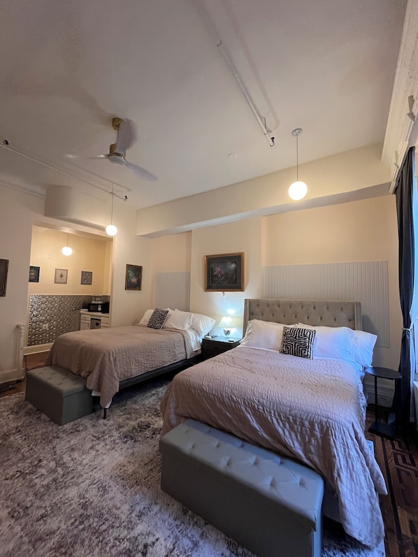 A hotel room with two beds, a sitting area, a vanity, and a ceiling fan.