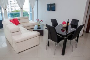 Ein modernes Wohnzimmer mit einer Couch, einem Esstisch mit Stühlen, einem an der Wand befestigten Fernseher und einem Balkon mit Blick auf Grünflächen.