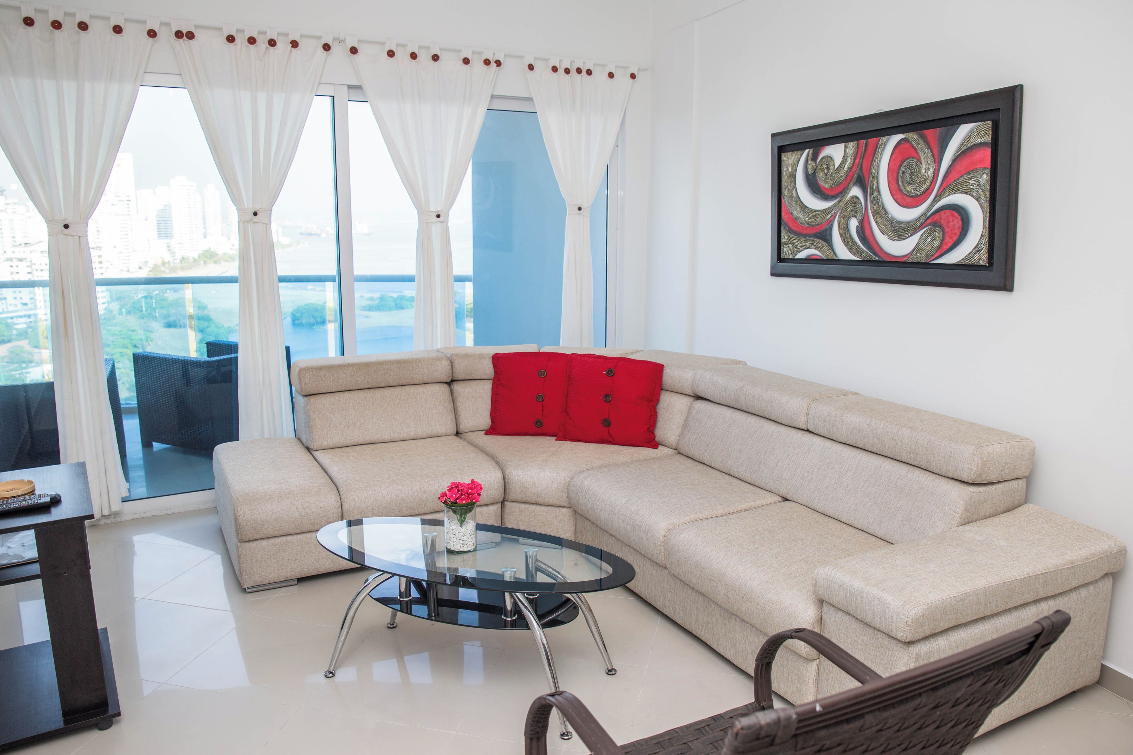 Una sala de estar con un sofá seccional color beige, una mesa de centro de vidrio con un florero, un sillón de mimbre y un cuadro abstracto enmarcado en la pared.