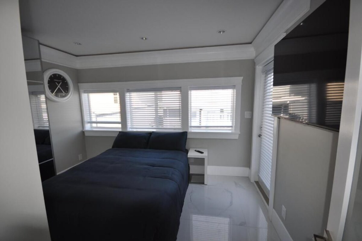 A bedroom with a bed, a nightstand, a clock, and a window with blinds.