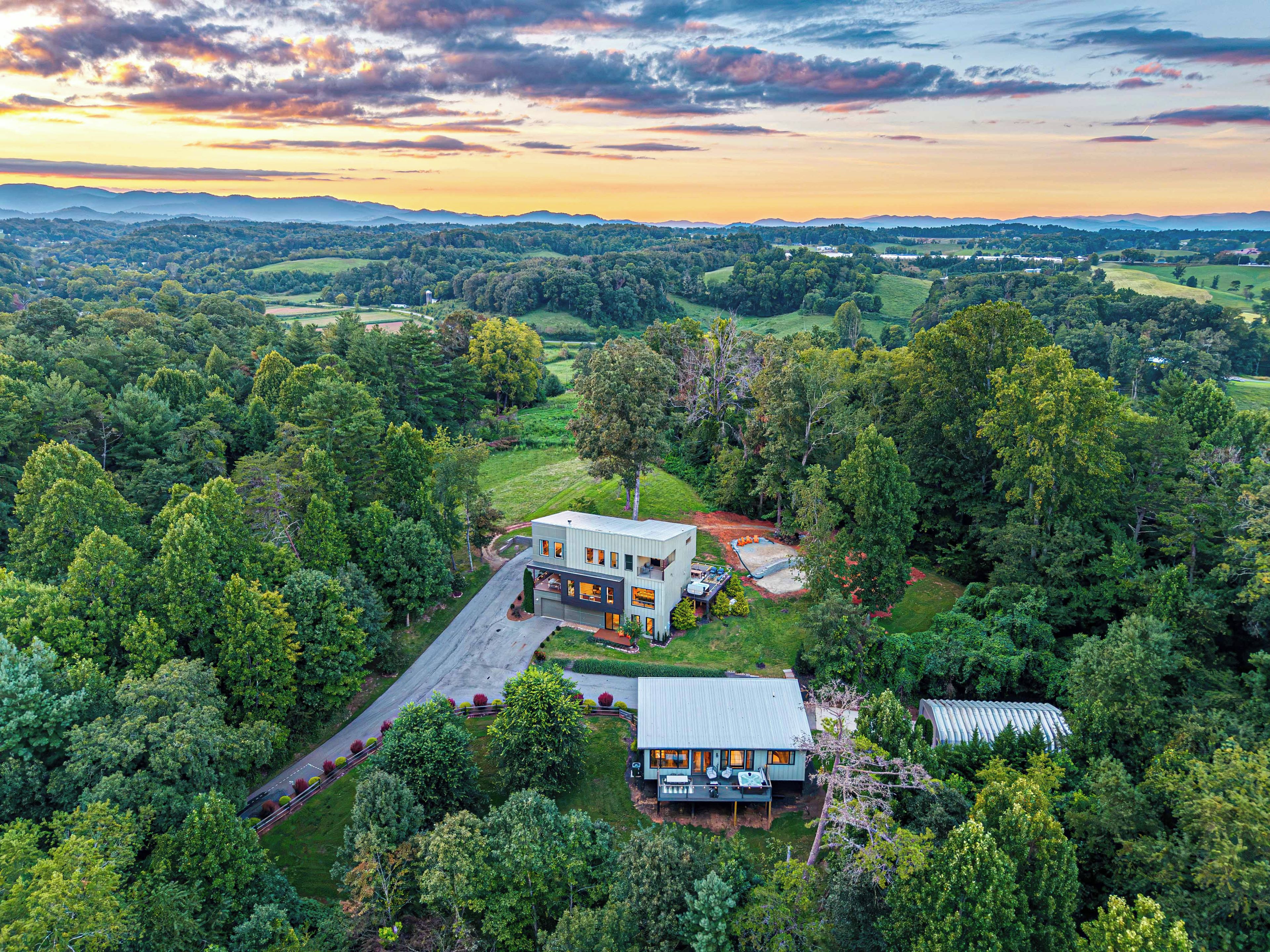 A modern house surrounded by dense forest, with a large driveway and a separate building with a sloped roof.