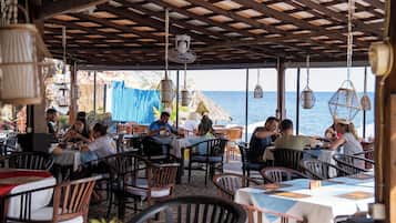 An outdoor dining area with tables and chairs, a thatched-roof structure, hanging lanterns, and a view of the sea.