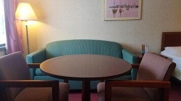 Una habitación de hotel con una mesa redonda, dos sillones, una cama, una lámpara y un cuadro en la pared.
