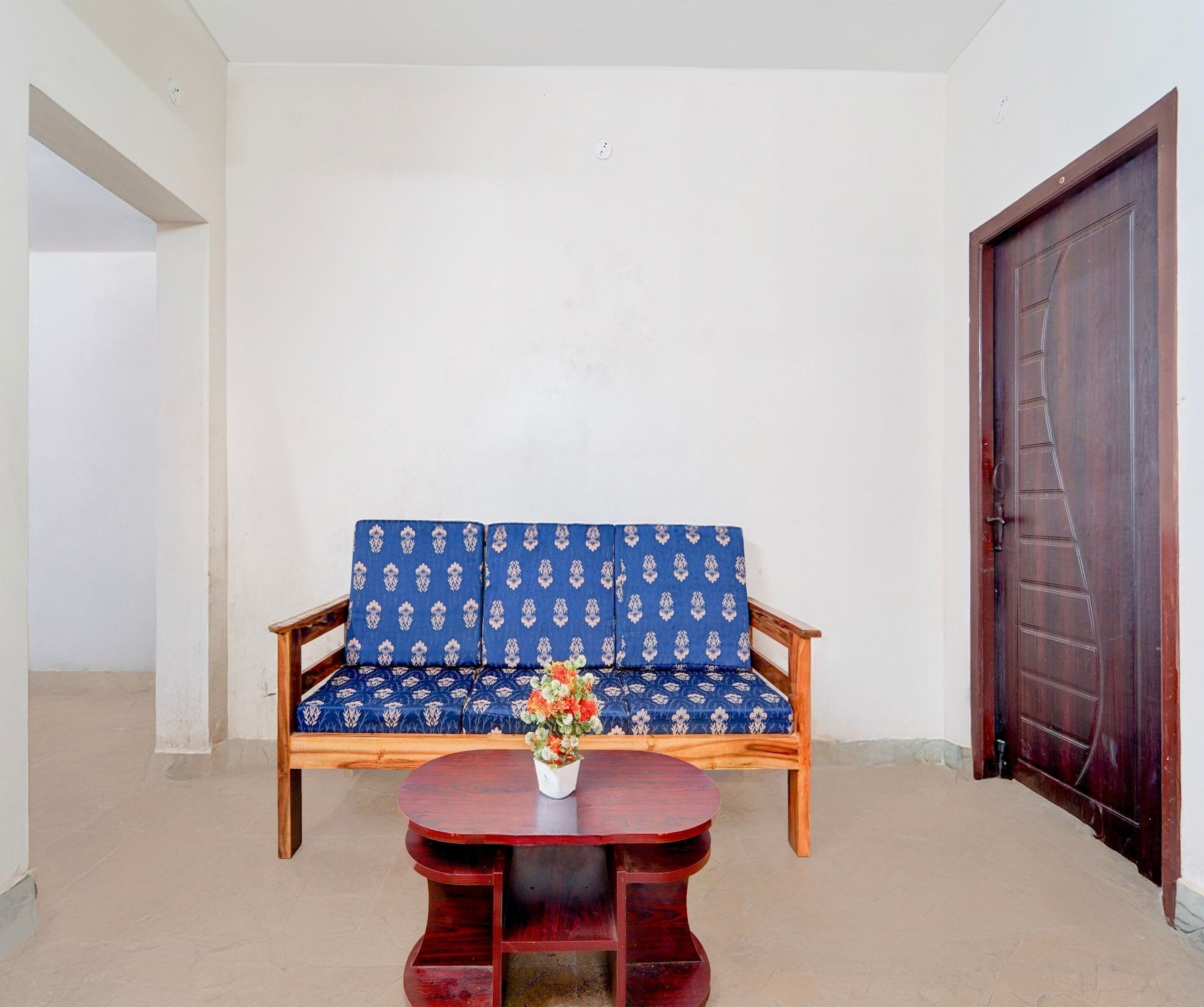 A room with a wooden sofa against a white wall, a wooden table with a vase of flowers, and a dark wooden door.