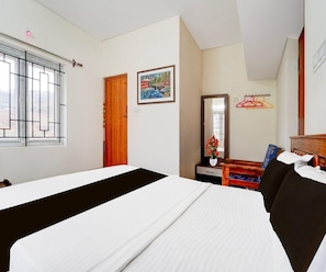 A hotel room with a bed, a wooden door, a painting, a mirror, a chair, and a window with bars. - Classic Aathira Hill Stay (Ootacamund)