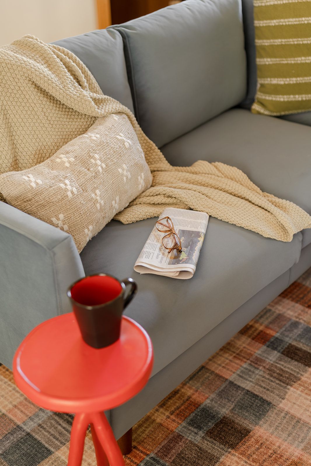 A grey sofa with a beige blanket and a folded newspaper with glasses on it.