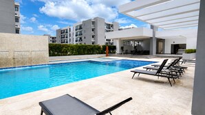 Una piscina con sillas de descanso, una zona de estar techada y edificios modernos al fondo.