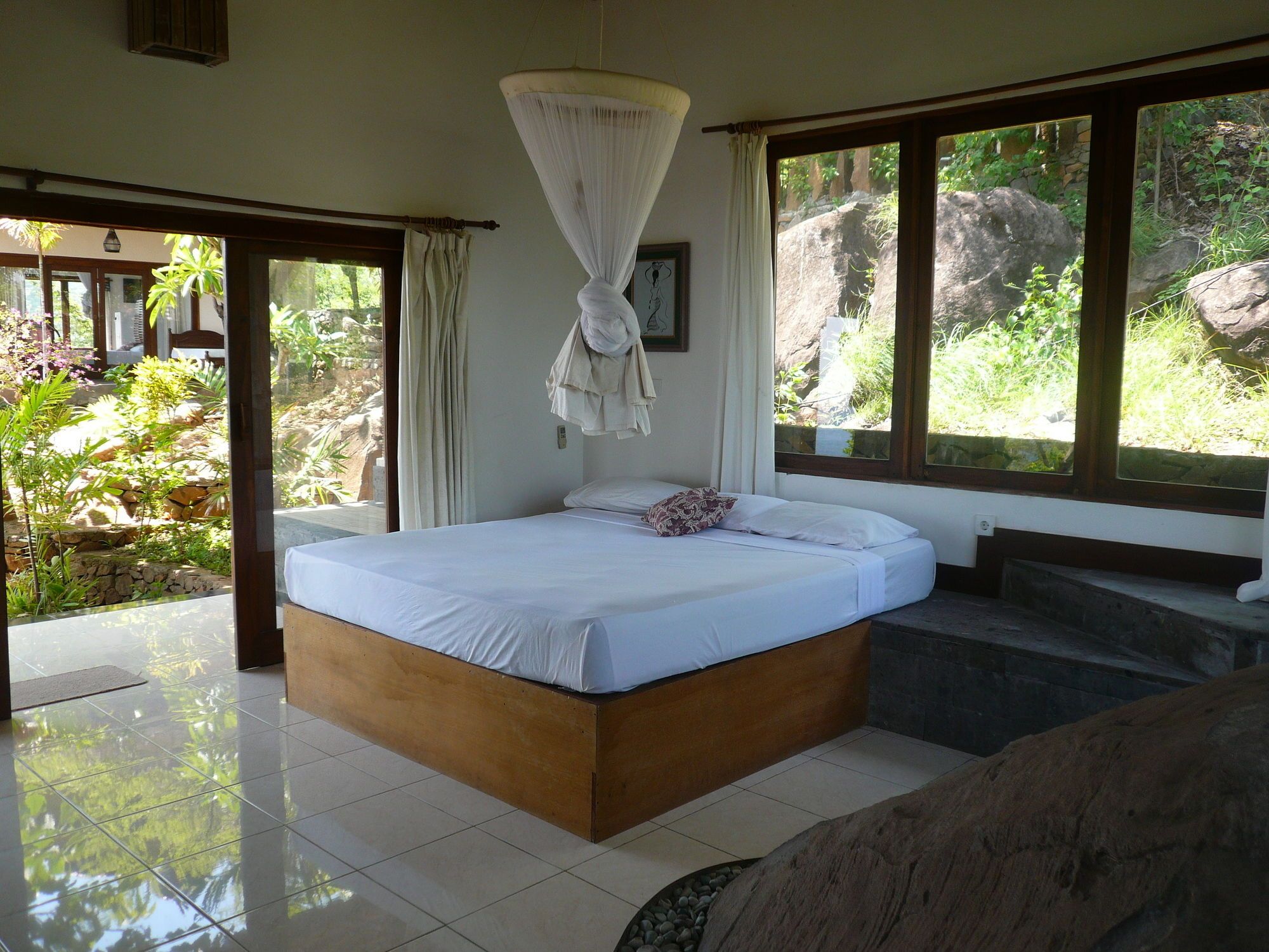A bedroom with a large bed, a hanging canopy, and a view of a garden through the window.