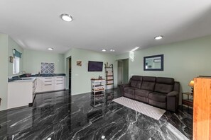 A modern living room with a black marble floor, a brown sofa, a TV mounted on the wall, and a kitchen area with white cabinets. - Hibisca 2BR Tropical Retreat with Pool & Outdoor Kitchen (Grote Berg)
