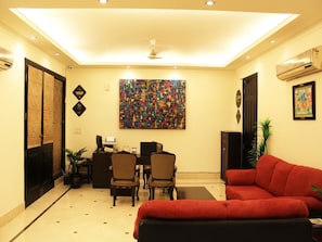 A room with a red sofa, a desk with a computer, a painting on the wall, and a ceiling fan. - OYO Premium Ambience Mall 2 (Gurugram)