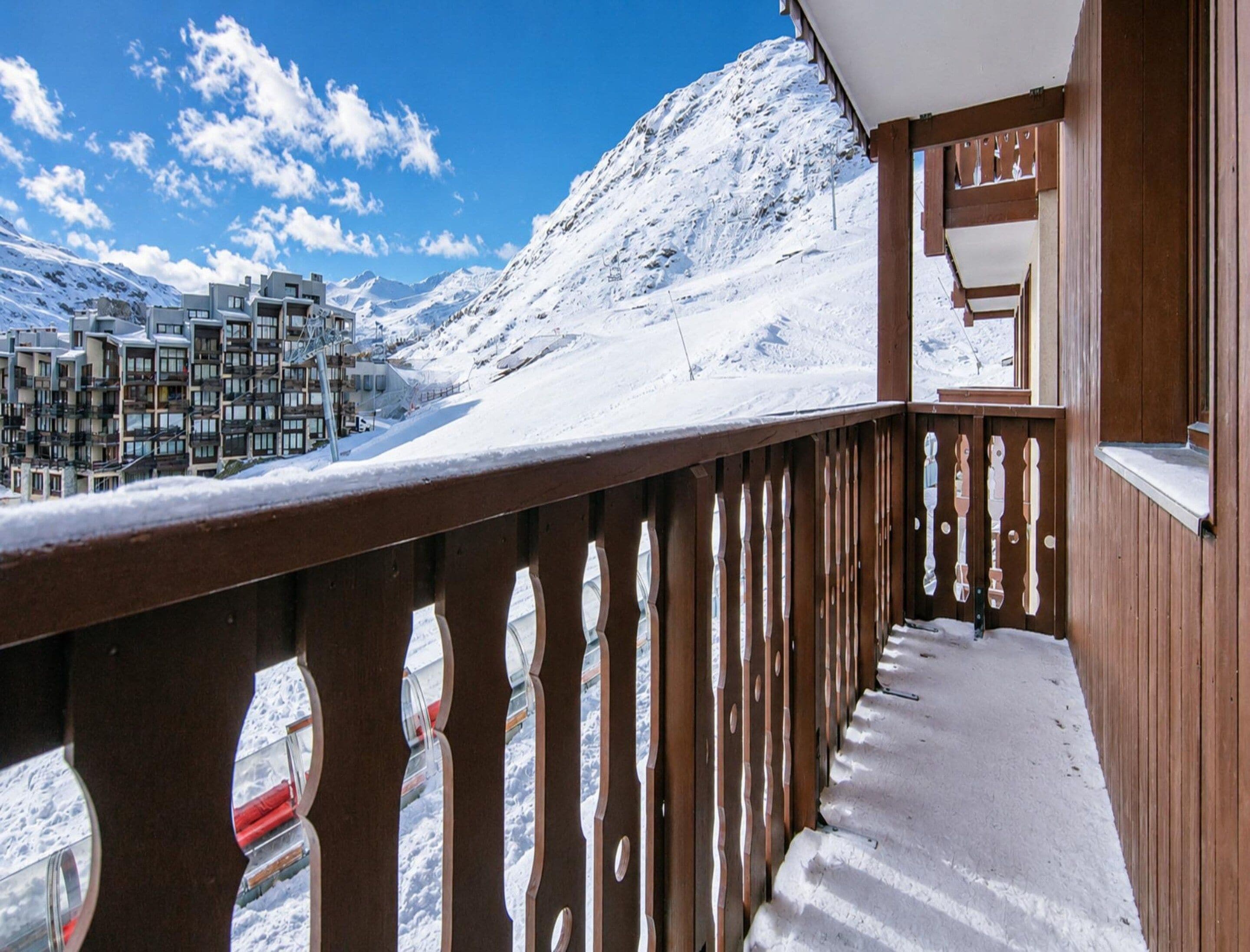 Un balcón de madera con suelo cubierto de nieve, con vistas a una zona montañosa con edificios.