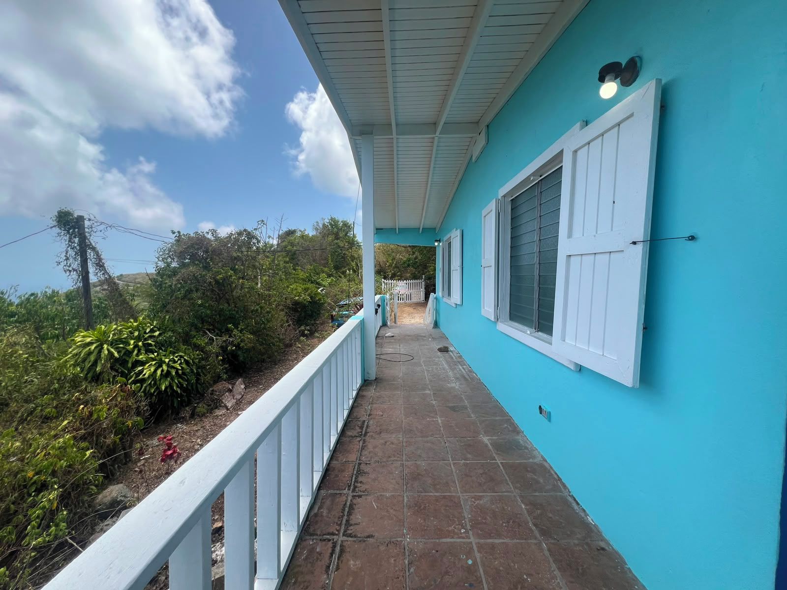 Une allée couverte, dont le sol est recouvert de carreaux, mène à un bâtiment doté de volets blancs et d’une façade de couleur turquoise.