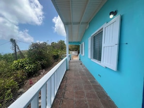 A covered walkway with a tiled floor leading to a building with white shutters and a teal exterior wall.
