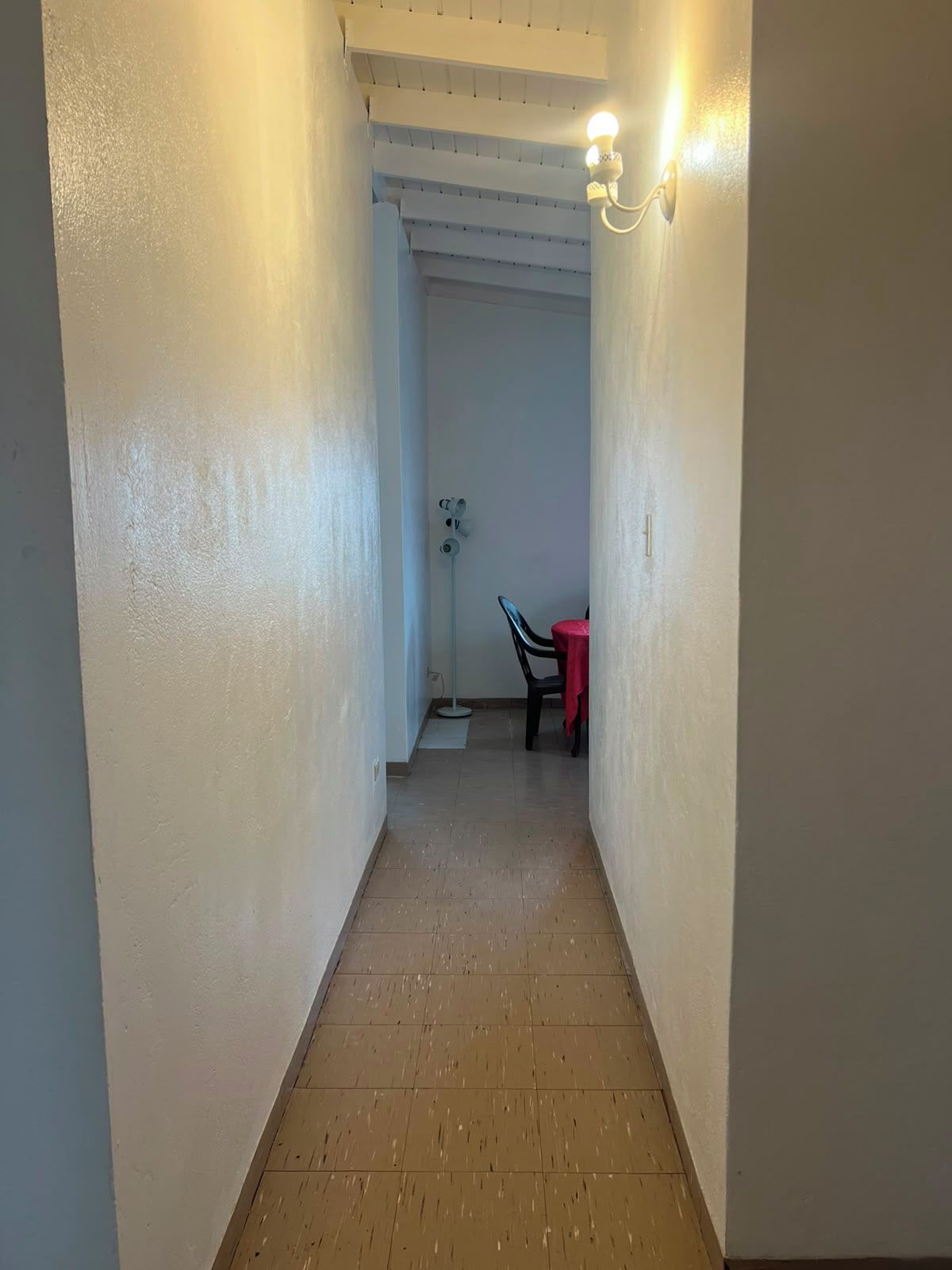 A narrow hallway with a light fixture on the ceiling and a chair with a red cover in the distance.