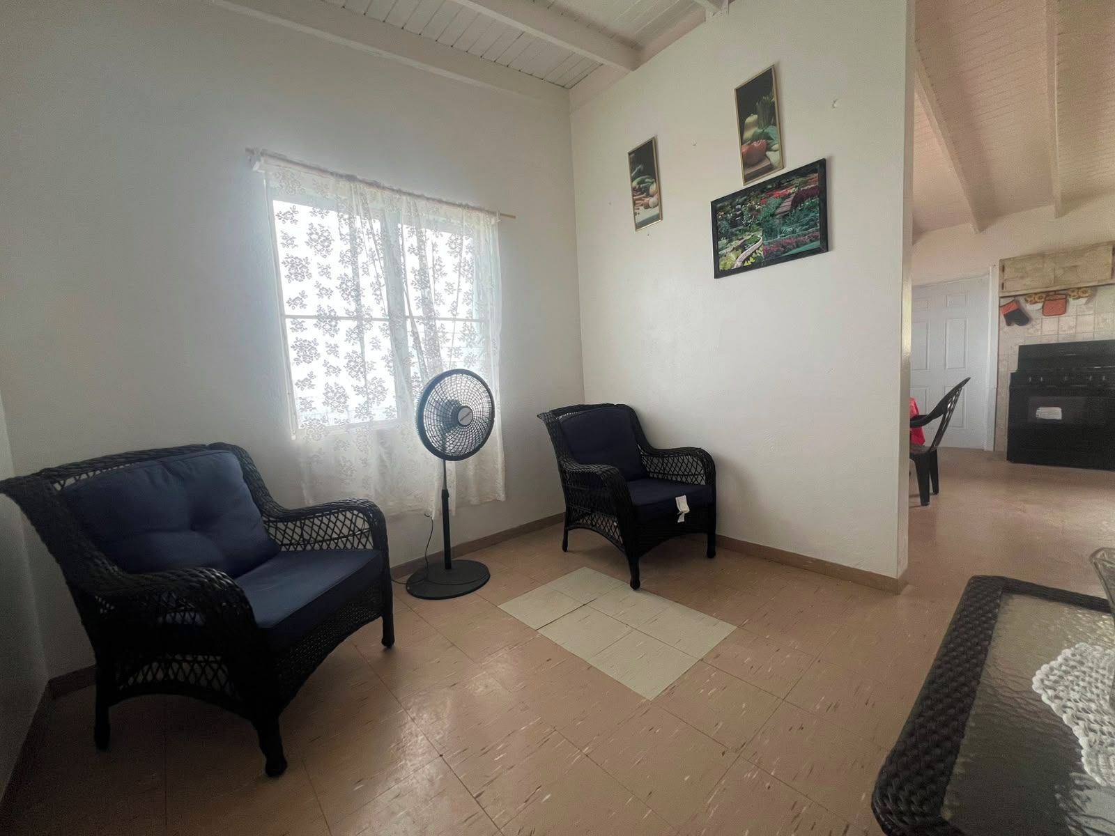 A living room with two black wicker chairs, a standing fan, and a window with lace curtains.