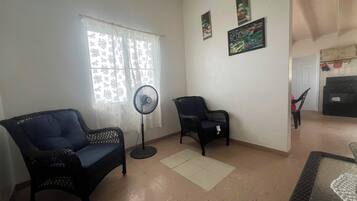 A living room with two black wicker chairs, a standing fan, and a window with lace curtains.