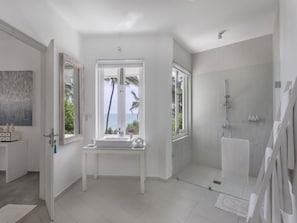 A modern bathroom with a shower, a vanity with a mirror, and a window providing a view of palm trees. - White Villa (Balapitiya)