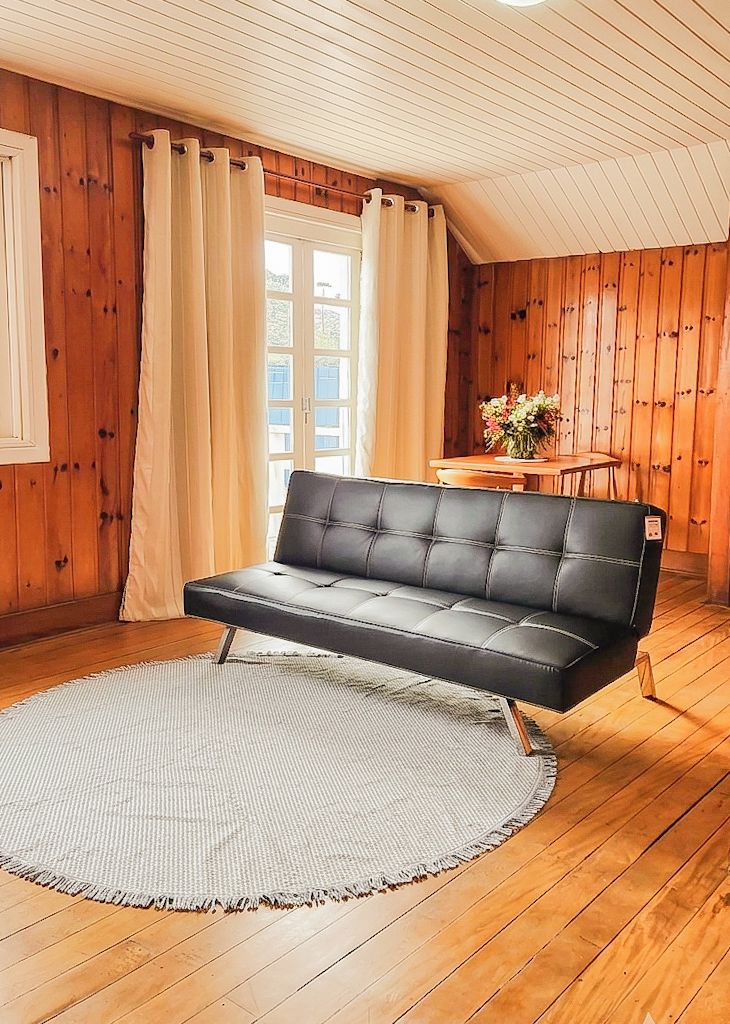 A room with wooden walls, a sofa, a round rug, a table with flowers, and a window with curtains.