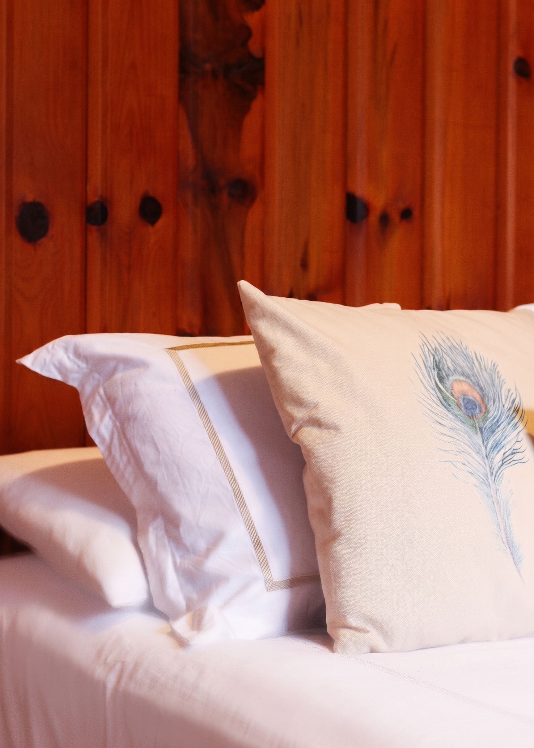 A bed with white bedding and two pillows, one plain and one with a peacock feather design.