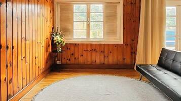 A room with wooden walls, a large window with shutters, a black leather sofa, and a small table with a vase.