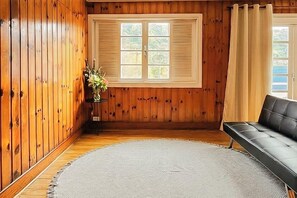 Una habitación con paredes de madera, un ventanal con contraventanas, un sofá de cuero negro y una pequeña mesa con un florero.