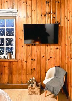 Una habitación con paneles de madera, un televisor de pantalla plana montado en la pared, una ventana con vistas, una silla y una cesta con flores.