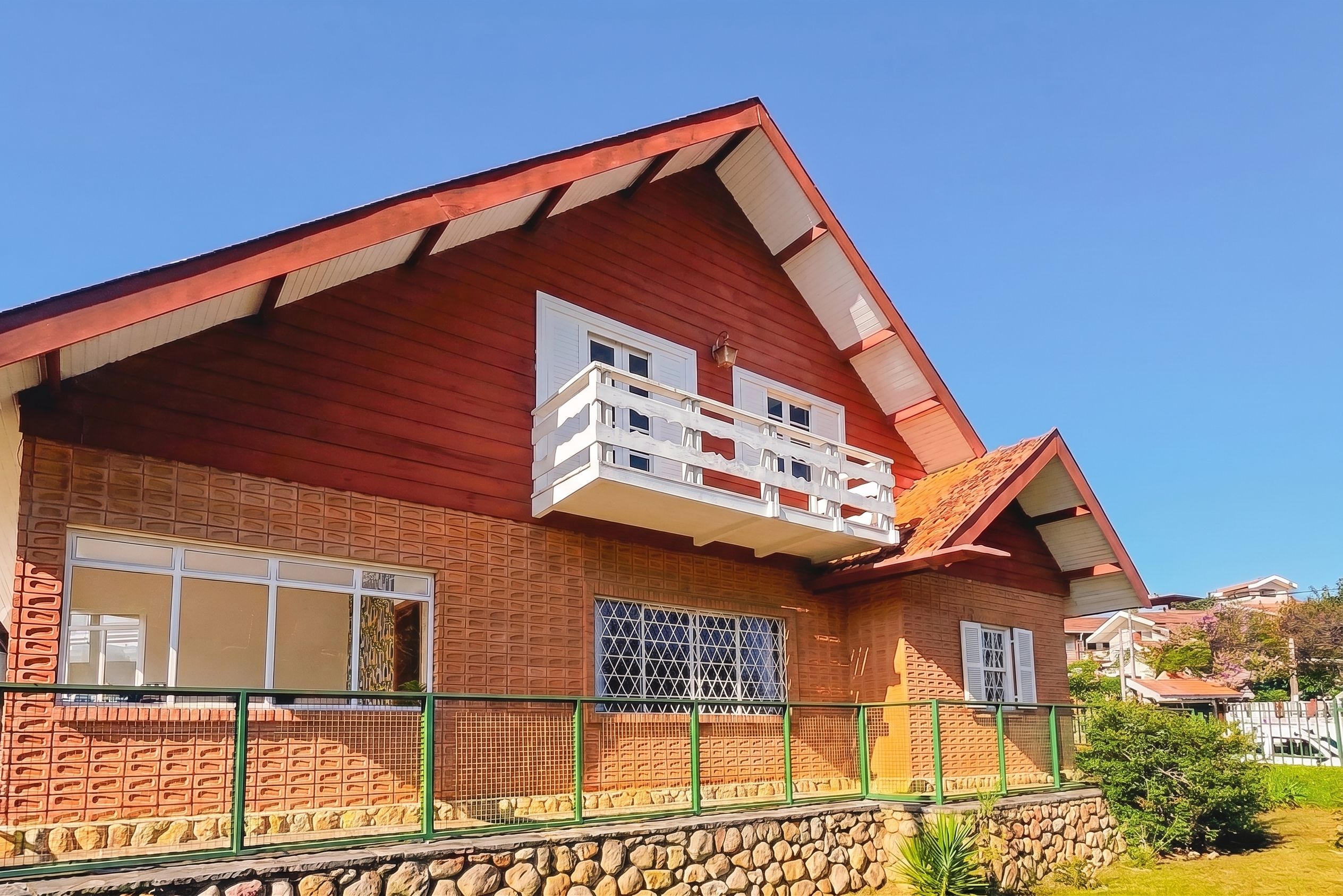 A two-story house with a red roof, a balcony on the second floor, and a stone foundation.