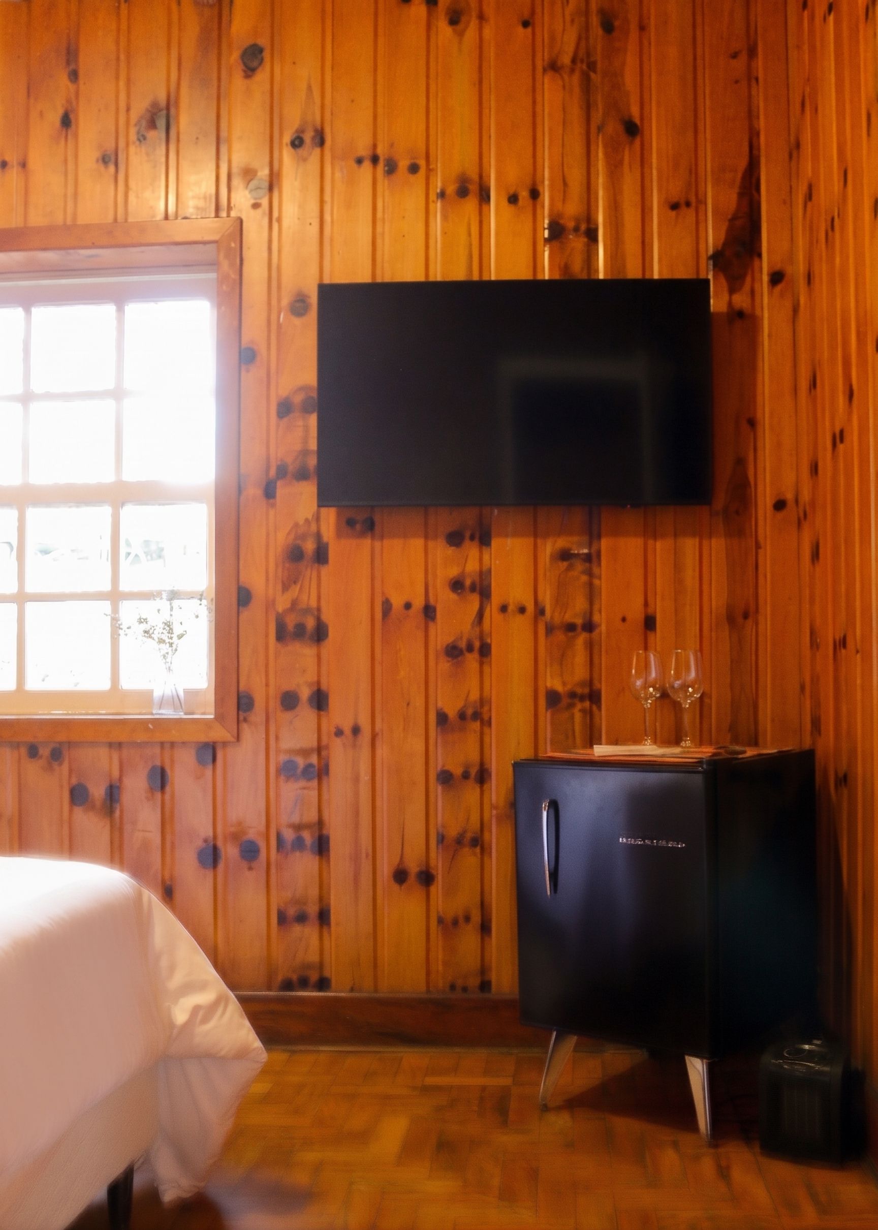A room with wooden walls, a bed with white bedding, a black entertainment unit with a flat-screen TV, and a window with a view.